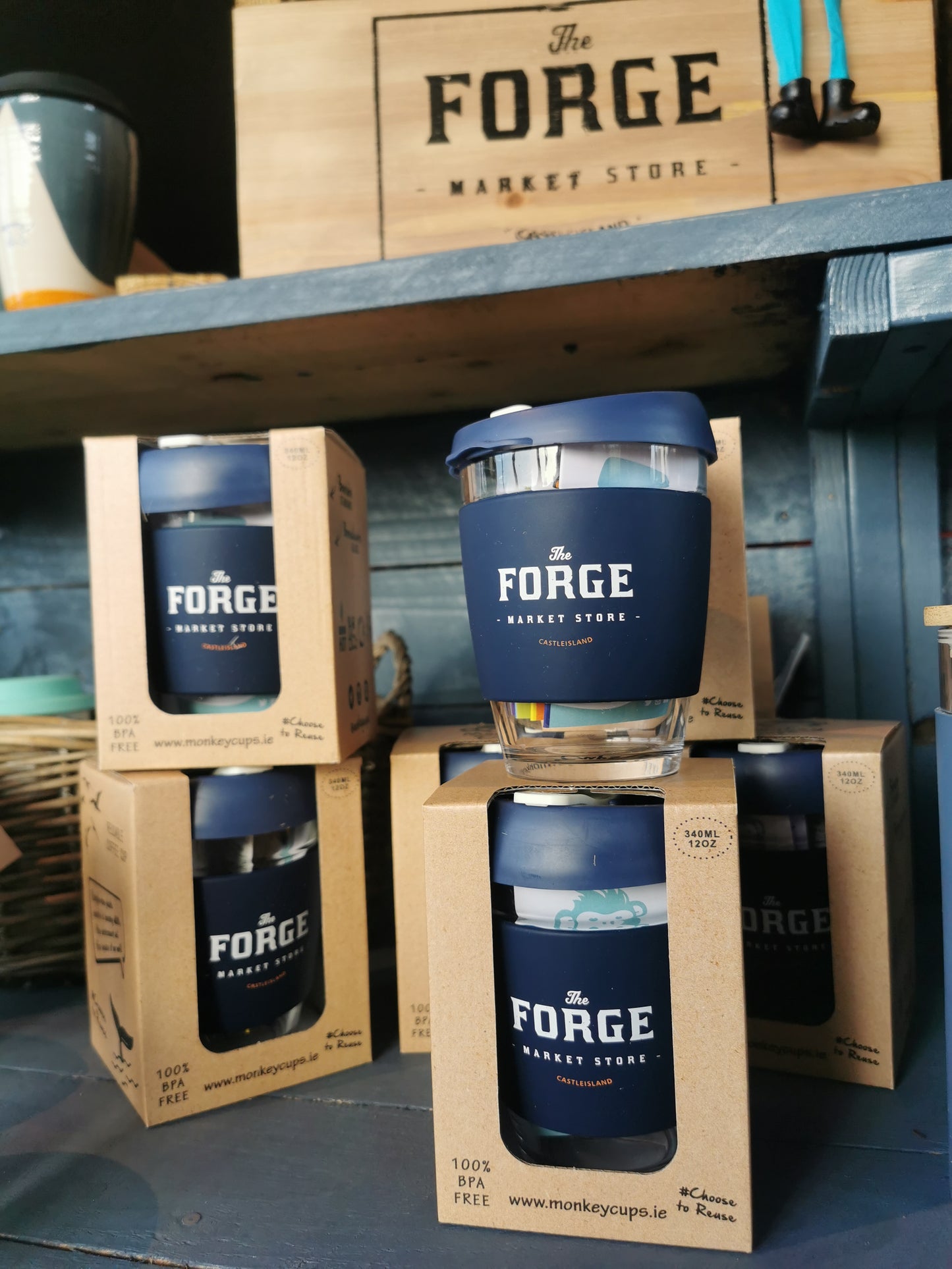 A display of Forge branded reusable glass coffee cups stacked in front of a wooden cabinet with the brand name Forge Market Store sign above.