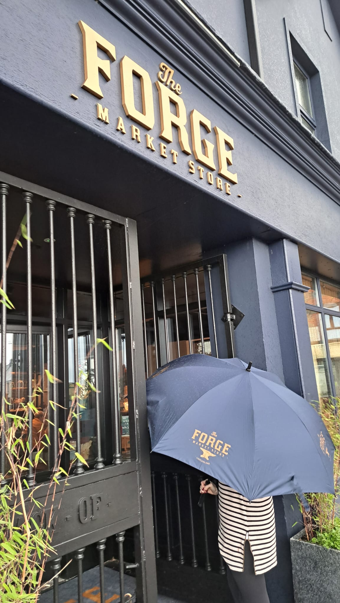 A person standing in front of a store named 'The Forge' using a navy blue umbrella with the word 'FORGE' printed on it.
