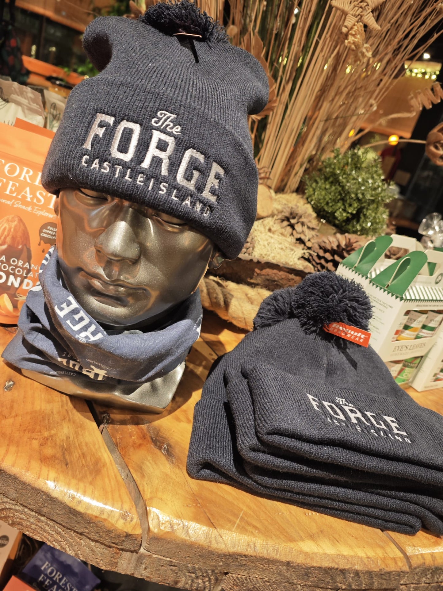 A black beanie with 'THE FORGE CASTLE ISLAND' text on the cuff, displayed on a wooden stand with a mannequin head wearing a similar hat in the background.