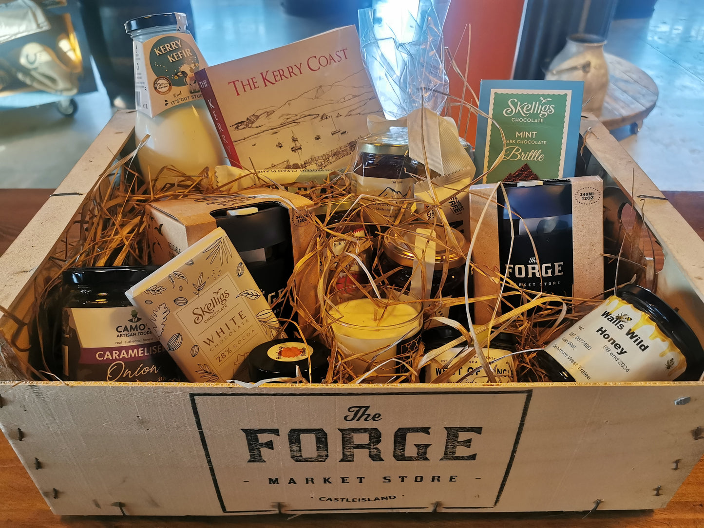 A wooden hamper basket containing a variety of items including bottles, a book, a card, and some foliage, all arranged on straw bedding.