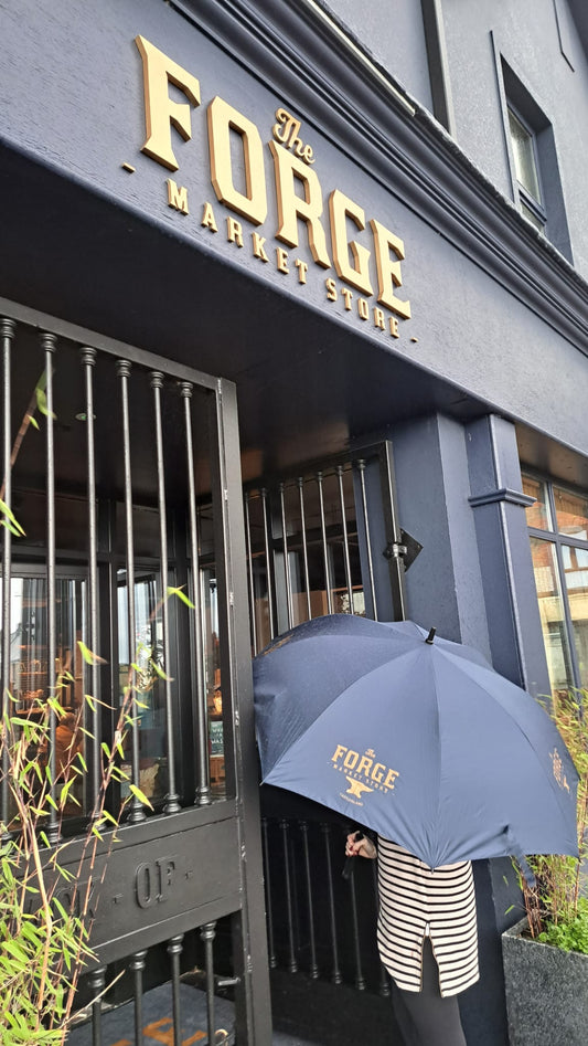 A person standing in front of a store named 'The Forge' using a navy blue umbrella with the word 'FORGE' printed on it.
